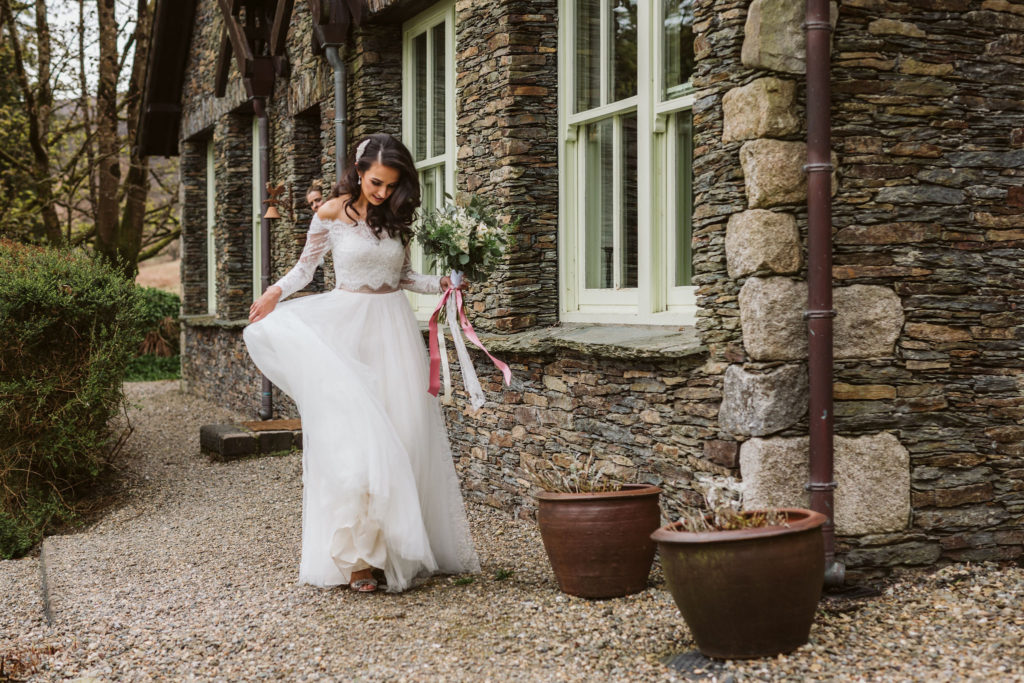 Real Wedding at Ballybeg House. Bride outside venue, she is holding her floral wedding bouquet and swishing her skirt. Her eyes are fixed on the ground.