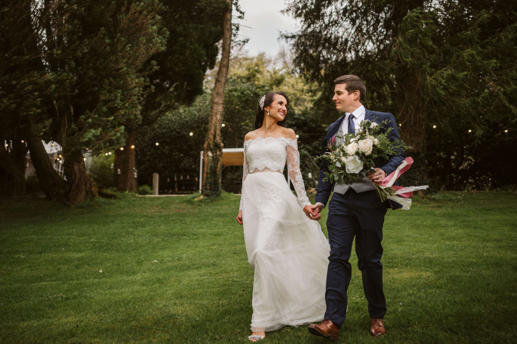 Real Wedding at Ballybeg House. Bride and GRoom walk hand in hand on the lawn outside the venue They are looking lovingly at each other. The Groom is holding the wedding bouquet in free hand. There are fairy lights visible in the background.
