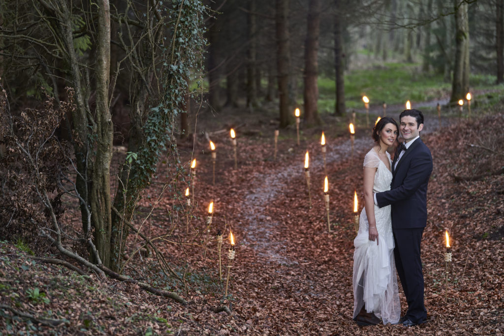 Bride and groom standing on leaf filled pathway. The leaves are brown and either side of the path is tree lines. There are lights lighting up the pathway. Location The Johnstown Estate.