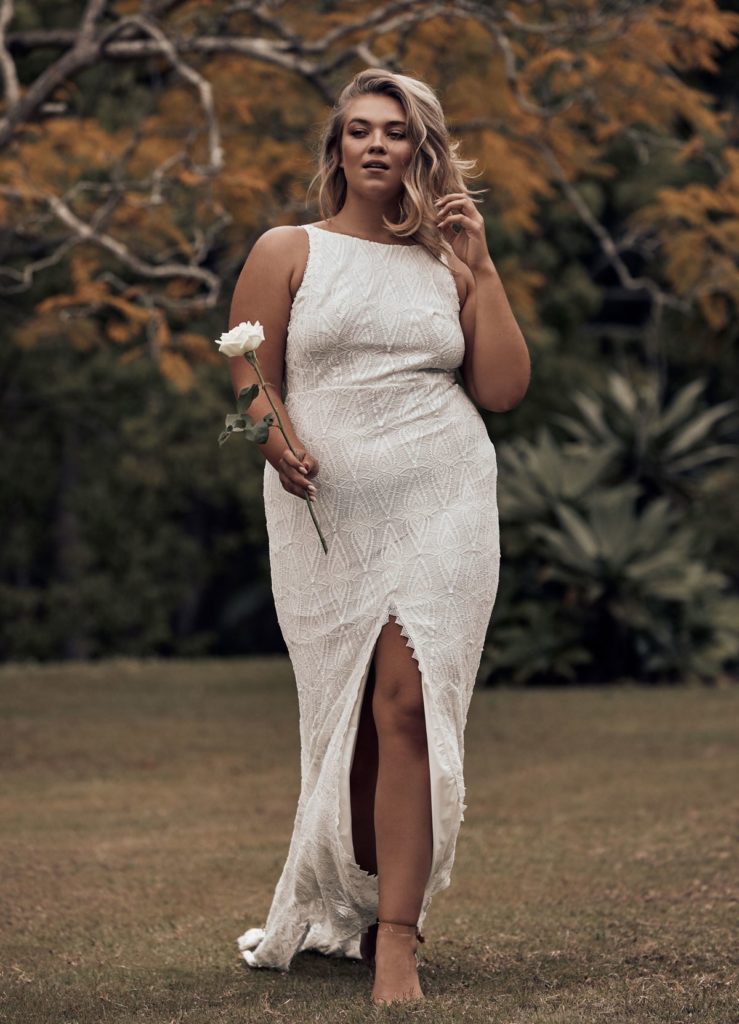 Curvy bride in a floor length, fitted white dress with a front slit to mid thigh and sport cut neckline