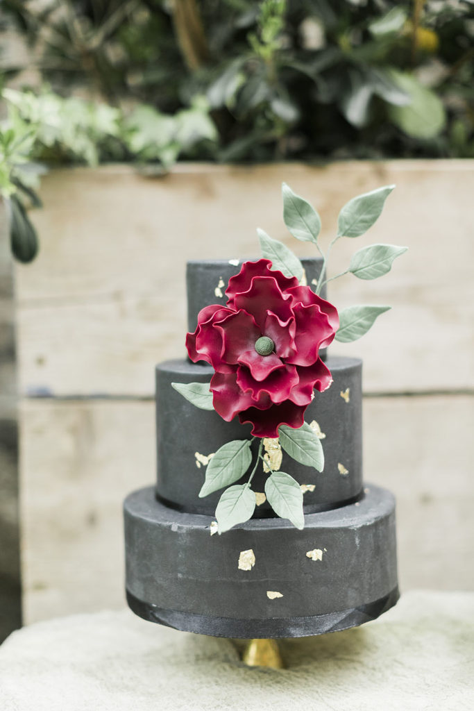 Black fondant, three tier wedding cake with large red fondant flower, fondant greenery and gold leaf decorating the front of the cake