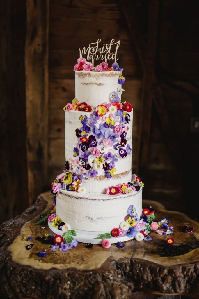 Three tier wedding cake decorated with rustic style white buttercream icing, a "Just Married" cake topper and adorned with purple, pink, yellow and white edible flowers across all three layers