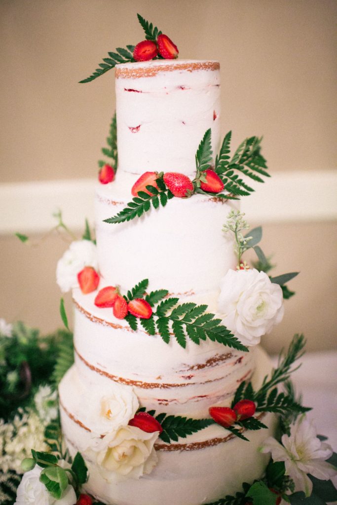 Four tier wedding cake decorated with rustic style white buttercream icing, ferns, white roses and sliced strawberry 