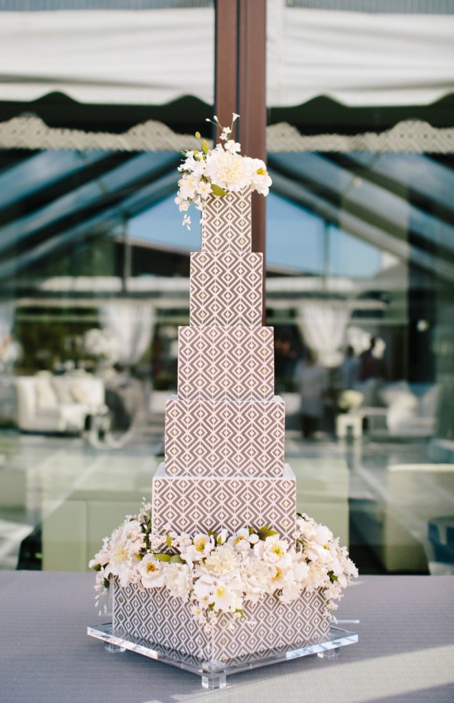 A six tier grey and white geometric designed wedding cake adorned with while flowers on the top layer and the base layer