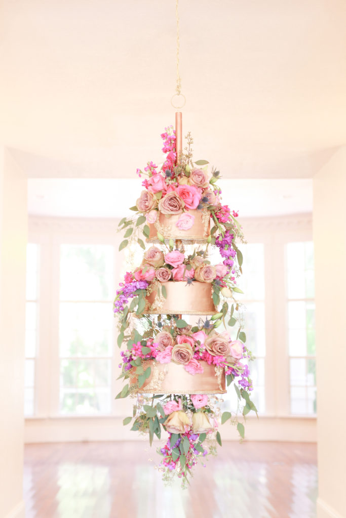 Three tiered gold wedding cake hanging from the ceiling covered in pink, purple and white roses with draping greenery