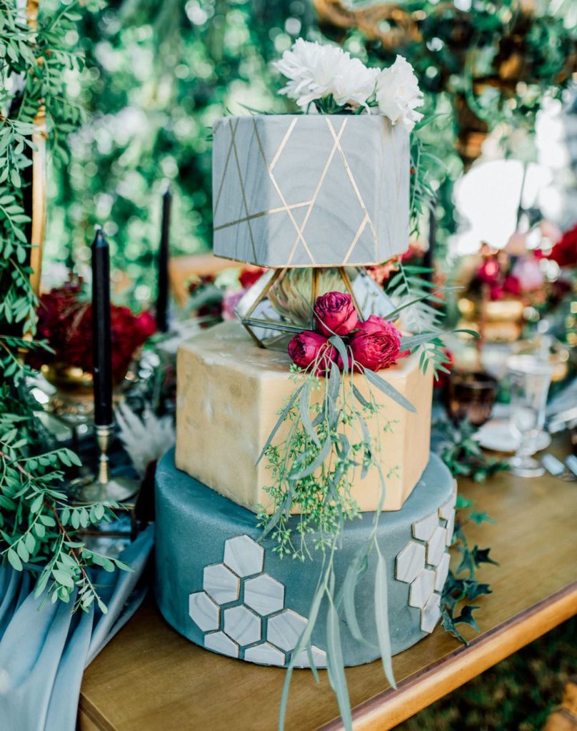 Three tier wedding cake with geometric spacer between top and middle layers. Bottom layer is green with grey hexagons, middle is gold and the top layer is covered in grey fondant with gold geometric lines