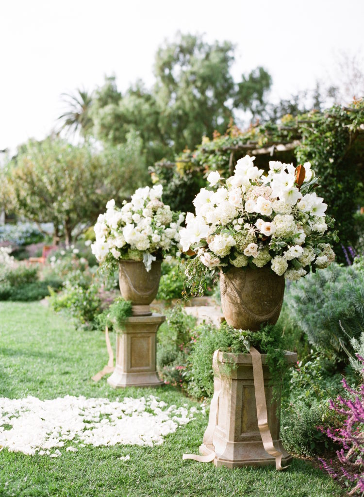 Two large stone vases full of large white flowers and greenery. At the base of the vases are beige ribbon and wrapped vines