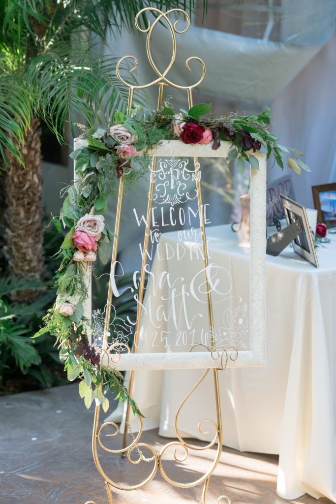 Glass pane in cream coloured frame with white calligraphy on it reading "Welcome to our wedding, Lisa & Matt" The frame sits on a gold stand and is decorated with a garland of greenery and pink, white and red flowers across the top, left hand side and half of the bottom