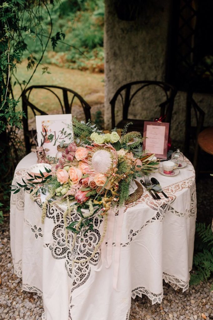 Large tied bouquet of assorted eucalyptus and grasses interspersed with pink roses and small white flowers on a small table covered in a lace white table cloth