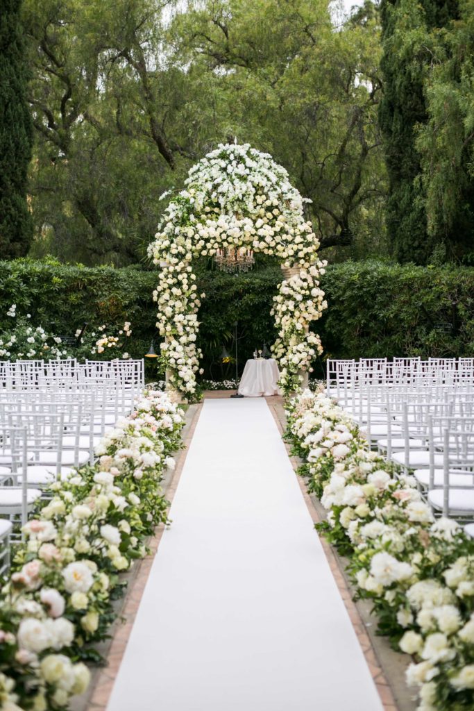 Large arch of white roses and greenery at the head of a white linen aisle bordered by white roses and greenery and white chairs