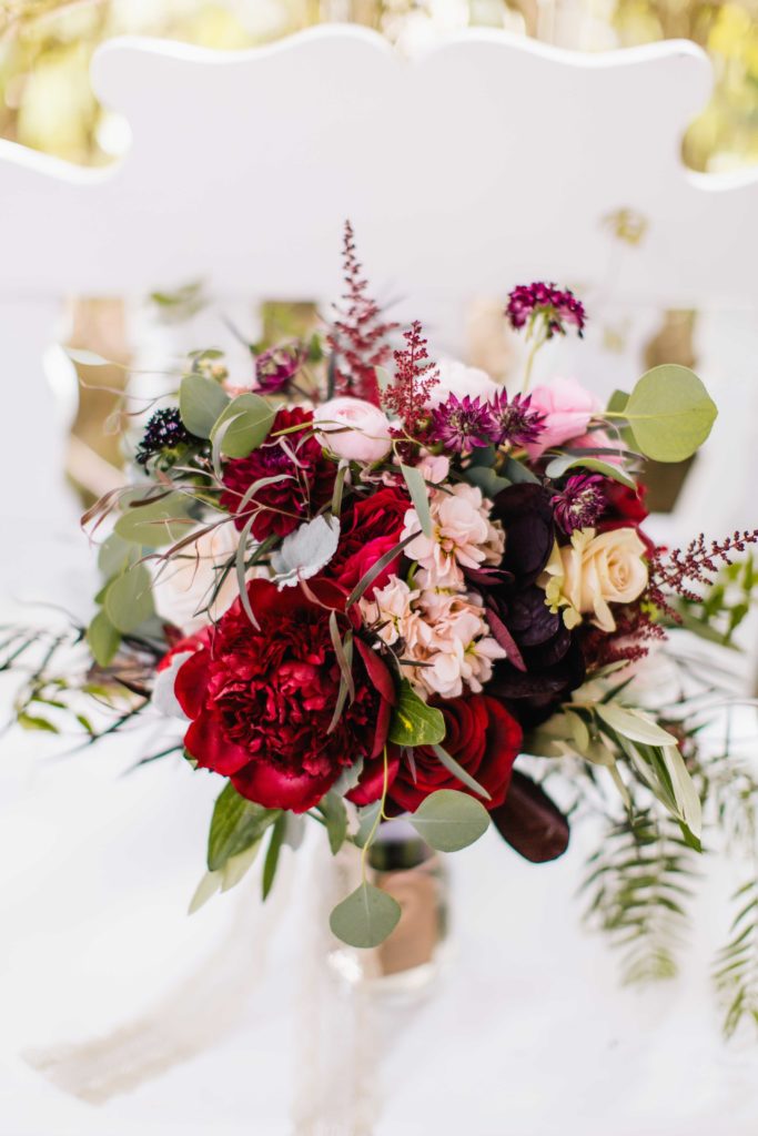 A tied bouquet of red, pink, white, purple and yellow flowers, green ferns and green leaves sitting on a white chair