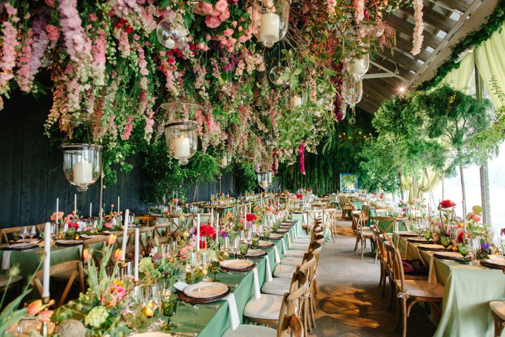 Large banquet tables with pink and green floral displays in the centre. A large mass of pink flowers, various greenery and lanterns hang from the ceiling above the tables