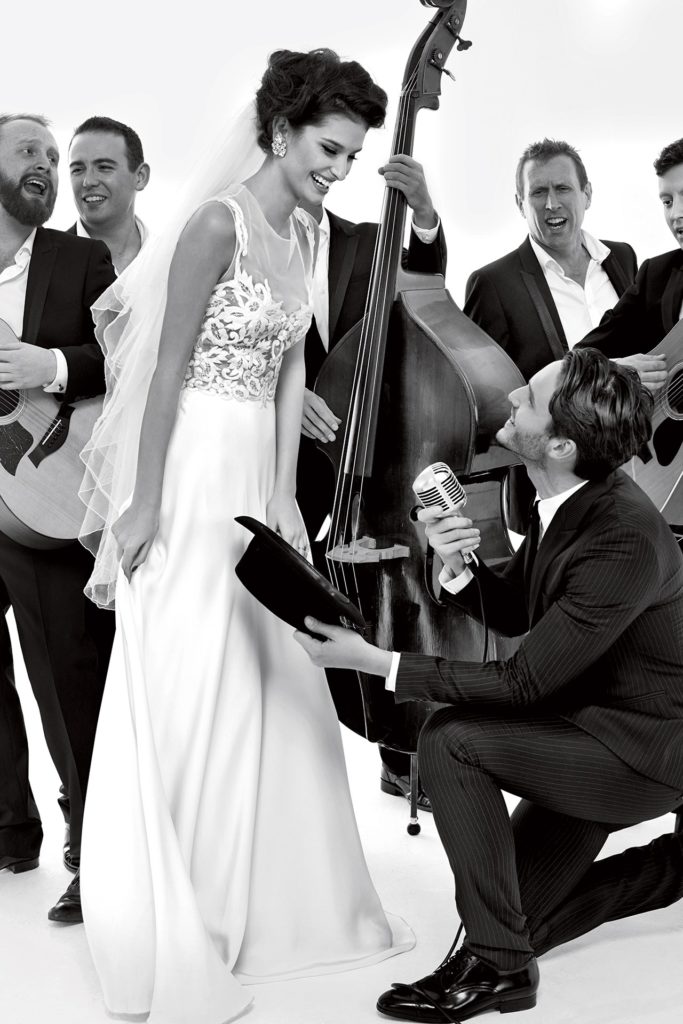 Bride in floor length, white dress stands in front of a band while the groom sings to her on one knee