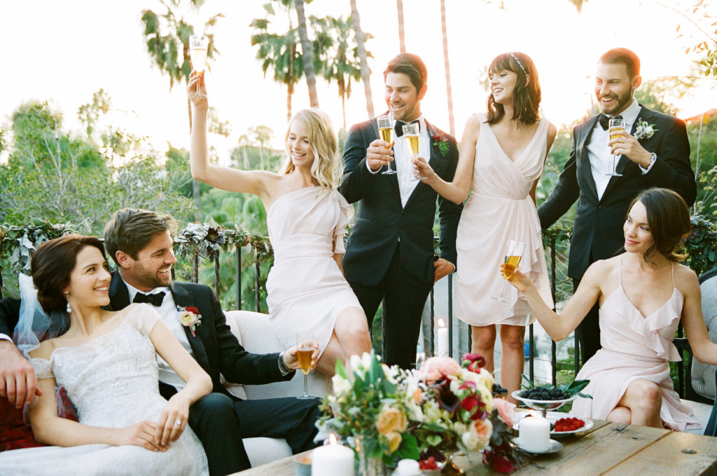 Five people in formal outfits toasting champagne to a seated bride and groom outside on a balcony 