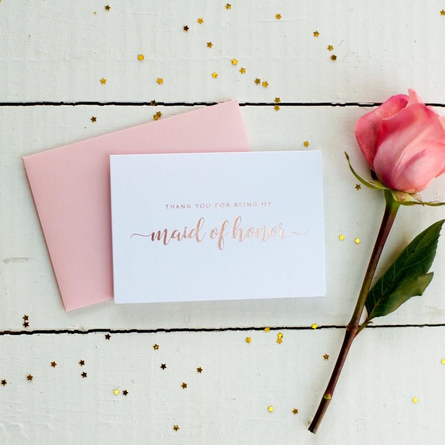 A white card with rose gold calligraphy stating "Thank you for being my maid of honor" sitting on top of a dusty pink envelope. Beside that is a pink rose, this is sitting on top of a white wooden table and scattered with gold star confetti 