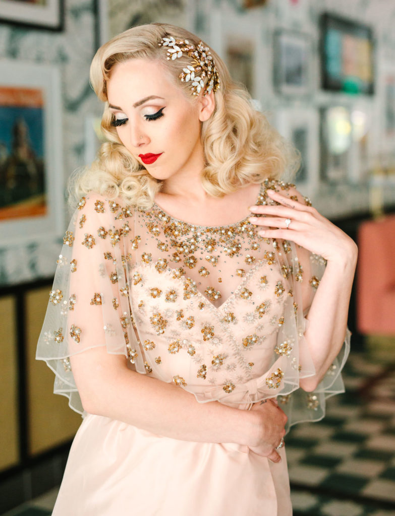 Blonde bride in champagne dress adorned with a sheer gold shawl covered in gold details. In her hair she has a large gold and pearl clip sweeping her curls into a structured vintage pin-curl hairdo