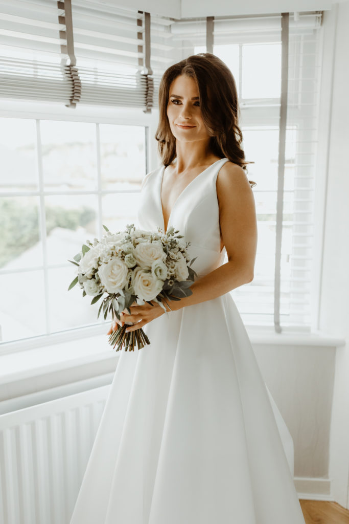 Irish bride standing next to a large bay window in a white satin Sassi Holford wedding dress