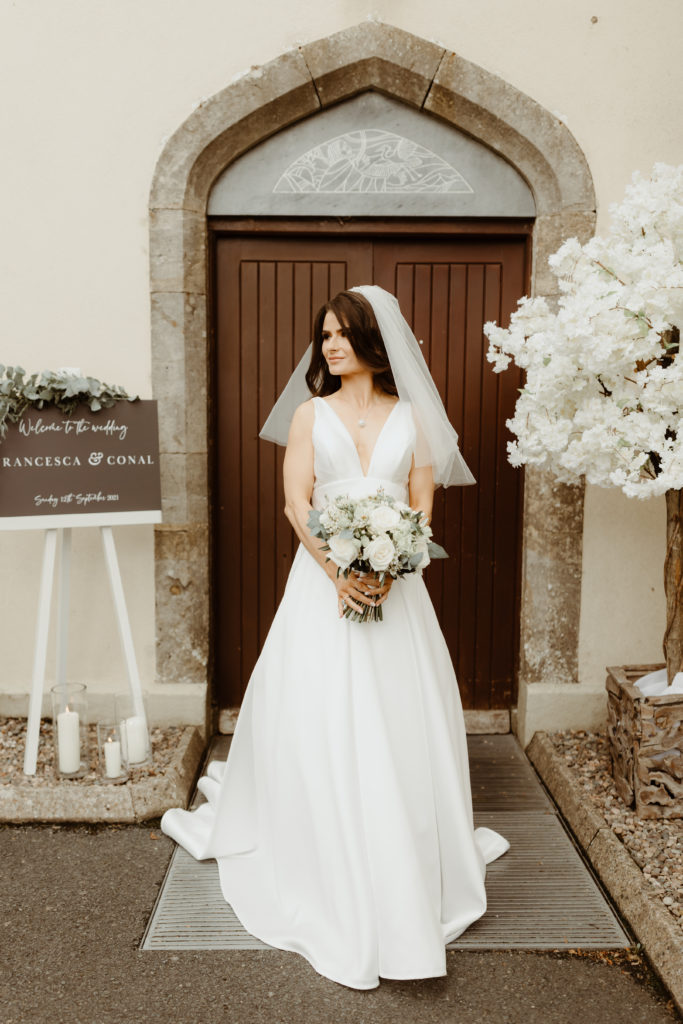 Irish bride standing next to a dark brown wooden church door in a white satin Sassi Holford wedding dress