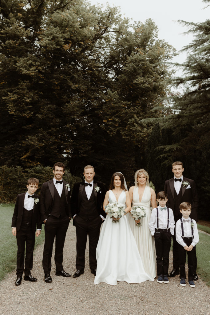 Irish bride and groom with bridesmaid in a long white dress, three groomsmen in black tuxedos and two small boys in white shirts with black suspenders standing in front of a group of trees