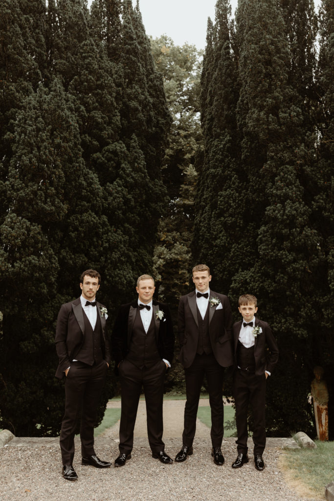 Irish groom with three groomsmen in black tuxedos and two small boys in white shirts with black suspenders standing in front of a group of trees