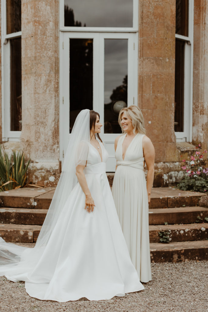 Irish bride with bridesmaid in a long white dress standing in front of a window in Castle Leslie