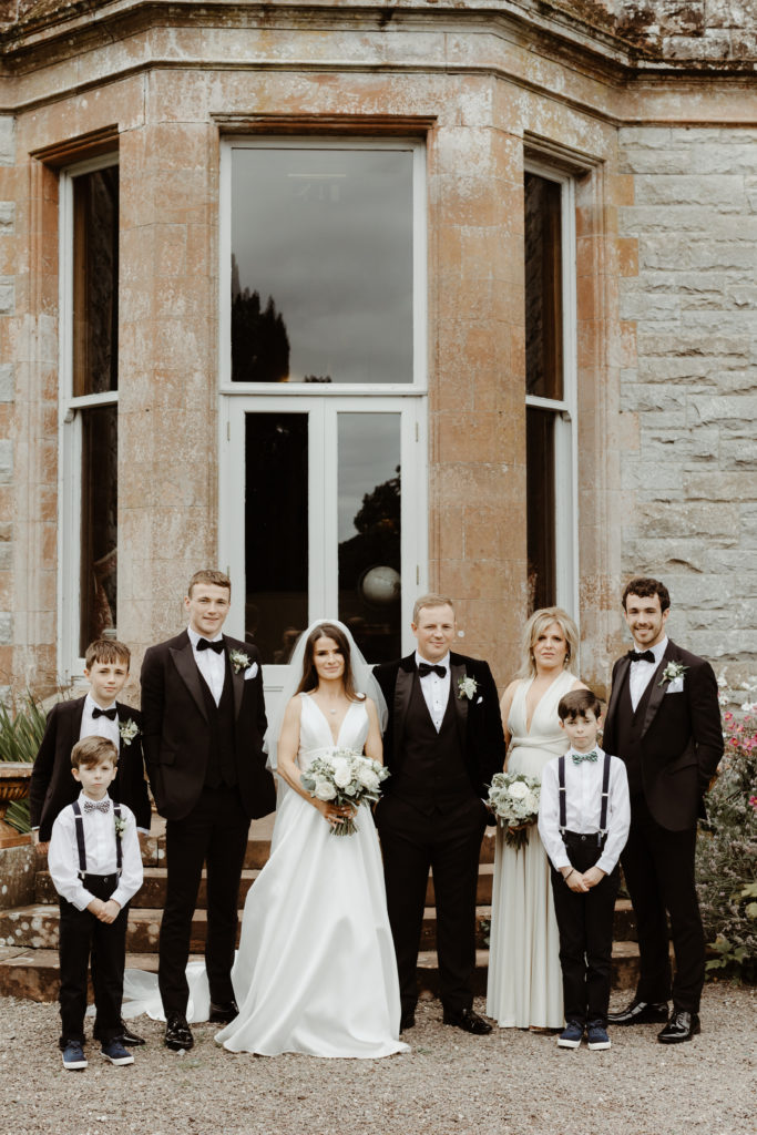 Irish bride and groom with bridesmaid in a long white dress, three groomsmen in black tuxedos and two small boys in white shirts with black suspenders standing in front of a window in Castle Leslie