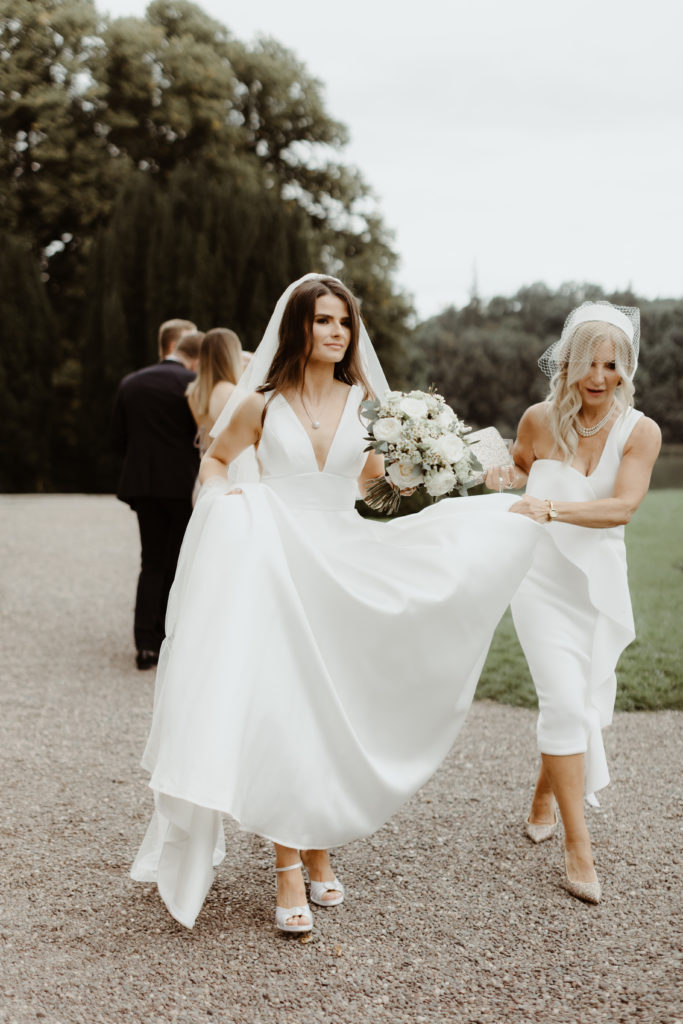 Irish bride in a white wedding dress and veil with and her mother in a long white dress walking from the car to the church while holding up her dress skirt
