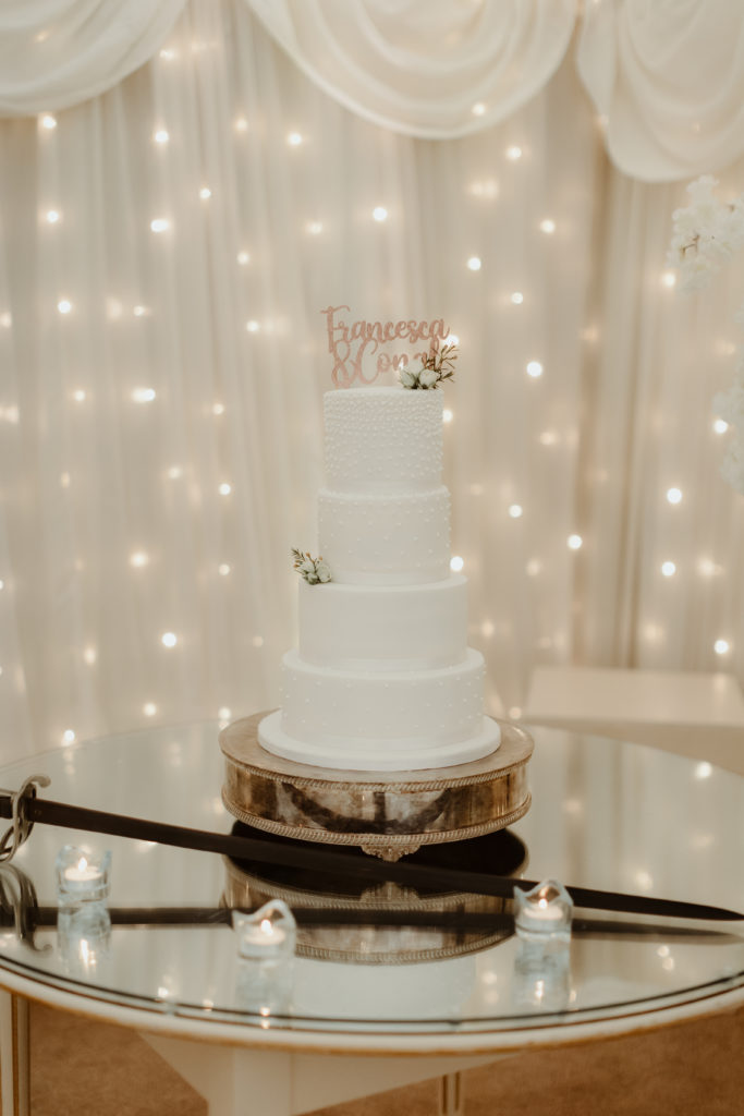 Four tier white wedding cake on a glass table with warm white string lights in the background