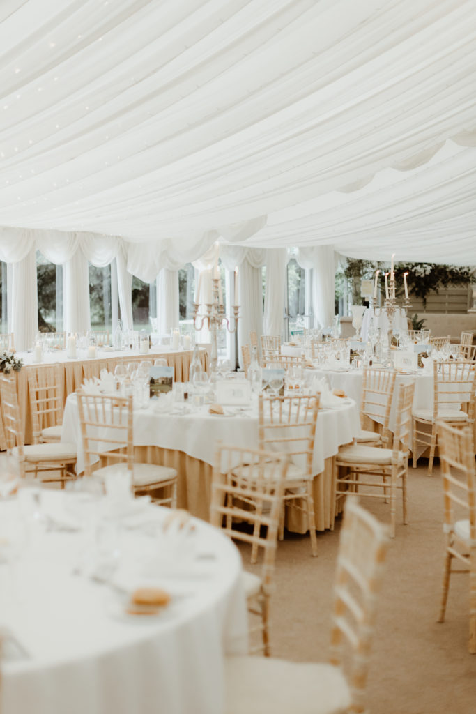 Inside of a large white marquee, a selection of brown tables and chairs draped with white fabric and covered with tableware